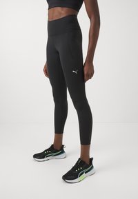 Czarne legginsy z wysokim stanem o dopasowanym kroju i boczną kieszenią, zestawione z czarnymi butami sportowymi z zielonymi akcentami i eleganckim wzornictwem.