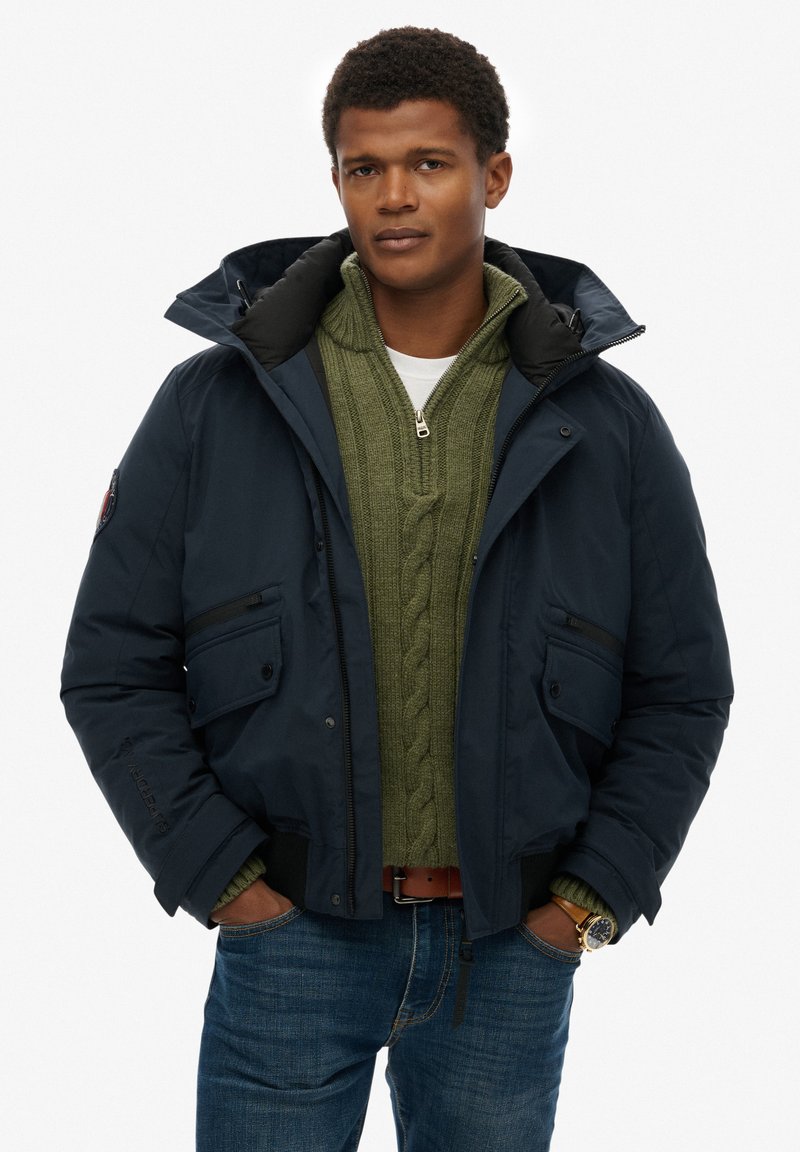 Granatowa ocieplana kurtka z kapturem, z dwoma przednimi kieszeniami, noszona na zielonym swetrze w warkocze i niebieskich dżinsach. Widoczny brązowy pasek i zegarek.