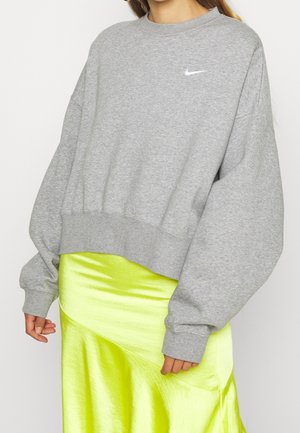 Gros plan d'une personne portant un sweat-shirt Nike gris oversize et une jupe en satin jaune fluo brillante.