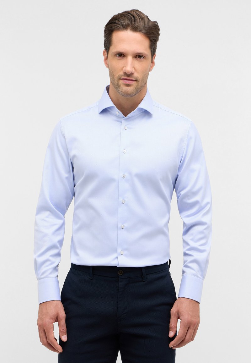 Camicia azzurro chiaro con maniche lunghe, colletto standard e bottoni bianchi. Indossata con pantaloni scuri, realizzati in tessuto texturizzato.