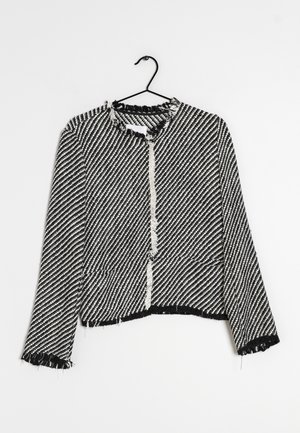 Veste texturée à rayures noires et blanches avec bords effilochés sur le col, les poignets et l'ourlet, suspendue à un cintre noir sur un fond blanc.