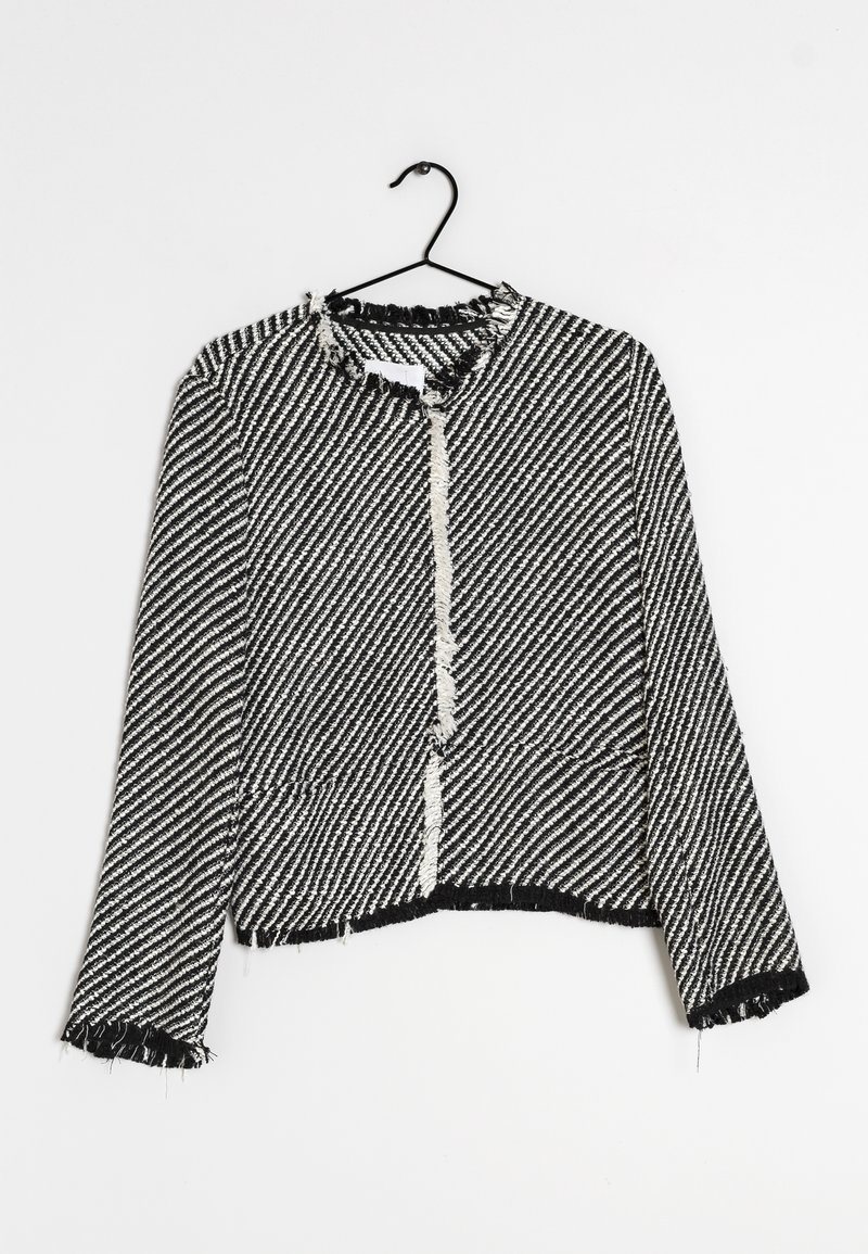 Veste texturée à rayures noires et blanches avec bords effilochés sur le col, les poignets et l'ourlet, suspendue à un cintre noir sur un fond blanc.
