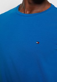 T-shirt en coton bleu royal avec un col rond. Présente un petit logo brodé en rouge, blanc et noir sur la poitrine. Texture lisse.