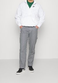 Vit Nike vindjacka med krage med kvartslång dragkedja, tillsammans med randiga grå byxor och svarta sneakers med en vit logga.