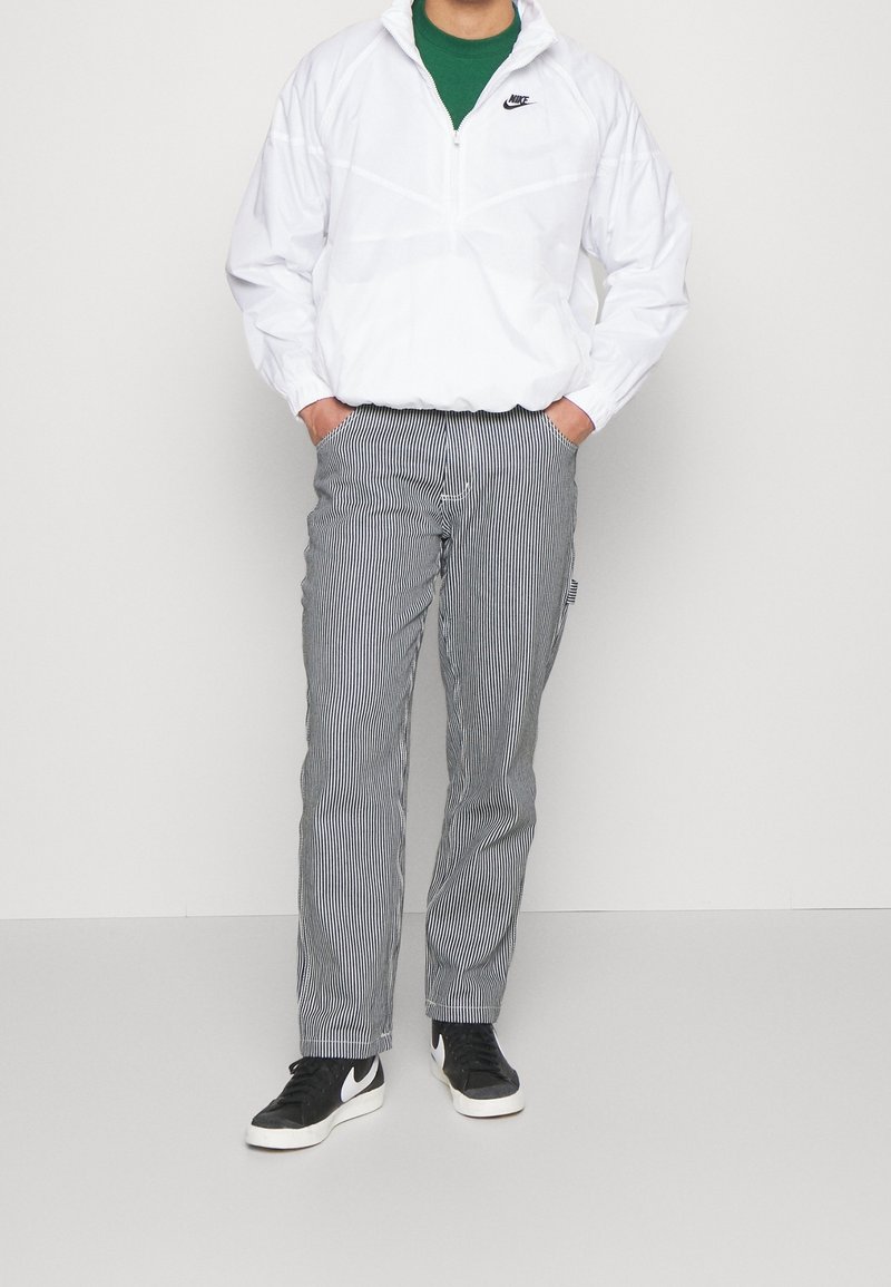 Vit Nike vindjacka med krage med kvartslång dragkedja, tillsammans med randiga grå byxor och svarta sneakers med en vit logga.