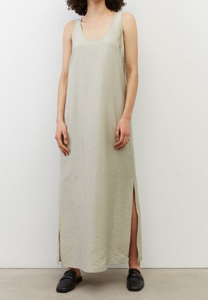 Femme portant une robe longue beige sans manches avec des fentes latérales, debout contre un mur blanc uni, associée à des chaussures noires sans lacets.