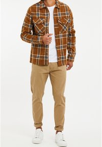 Mand iført brunt ternet skjorte over hvid t-shirt, tan bukser rullet op ved anklerne og hvide sneakers, stående mod hvid baggrund.