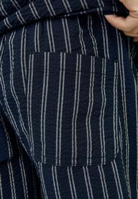 Tissu marine avec des rayures blanches, présentant un petit détail de poche. Le tissu a une finition texturée, et les rayures sont disposées verticalement.