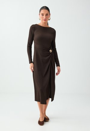 Femme portant une robe midi manches longues de couleur marron foncé avec un détail de boucle dorée à la taille, associée à des chaussures plates marron foncé et des boucles d’oreilles créoles.