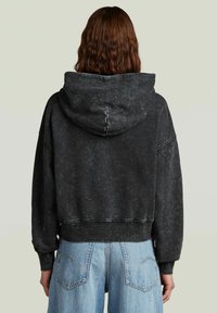 Felpa nera tagliata con una texture leggermente sbiadita, vestibilità oversize e cappuccio. Presenta polsini e orlo a coste. Abbinata a jeans azzurri chiari.
