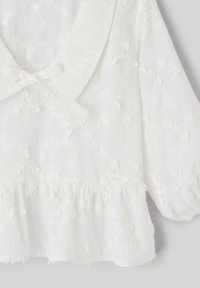 Blouse blanche à manches bouffantes, tissu embossé floral, grand col et détail de nœud noué à l'avant.