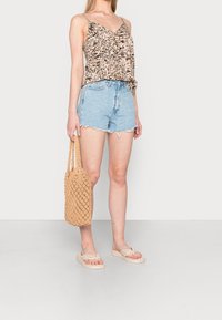 Top leggero a motivo, beige, con spalline sottili, abbinato a pantaloni corti in denim sfilacciati. Tiene una borsa intrecciata in tonalità beige e indossa infradito beige.