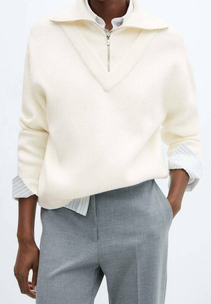Personne portant un pull côtelé crème avec une fermeture éclair à mi-hauteur sur une chemise blanche à fines rayures, associée à un pantalon gris ajusté.