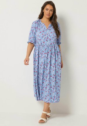 Kvinde med langt mørkt hår iført en lyseblå midi-kjole med rød og hvid blomsterprint og hvide sandaler, stående mod en ensfarvet baggrund.