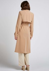 Manteau long beige avec une coupe décontractée, design croisé et ceinture à la taille. Tissu lisse avec des manches longues et une silhouette fluide.