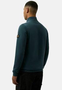 Turbia bluza z suwakiem i wysokim kołnierzem, z czarną naszywką logo na lewym rękawie oraz ściągaczami przy mankietach i u dołu. Prosty, jednolity design.