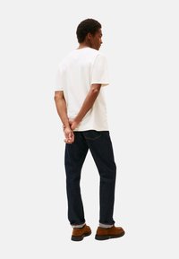 Hombre de pie con las manos entrelazadas detrás de la espalda, vistiendo una camiseta blanca, jeans oscuros y zapatos marrones, de espaldas frente a un fondo liso.