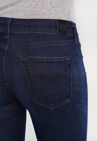 Vaqueros de mezclilla azul oscuro con una textura suave, que presentan un bolsillo trasero con un diseño de costura curva y una etiqueta negra en la cintura.