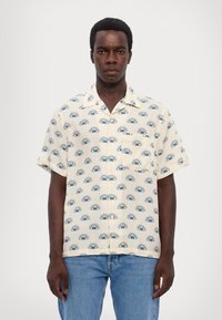 Korte mouwen, crème shirt met een herhalend blauw zon- en oogpatroon. Lichtgewicht stof, ontspannen pasvorm en een borstzakje.