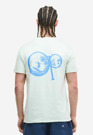 Man met gevlochten haar draagt een lichtgroen T-shirt met een blauwe surfscène afbeelding en marineblauwe shorts, van achteren op een witte achtergrond.