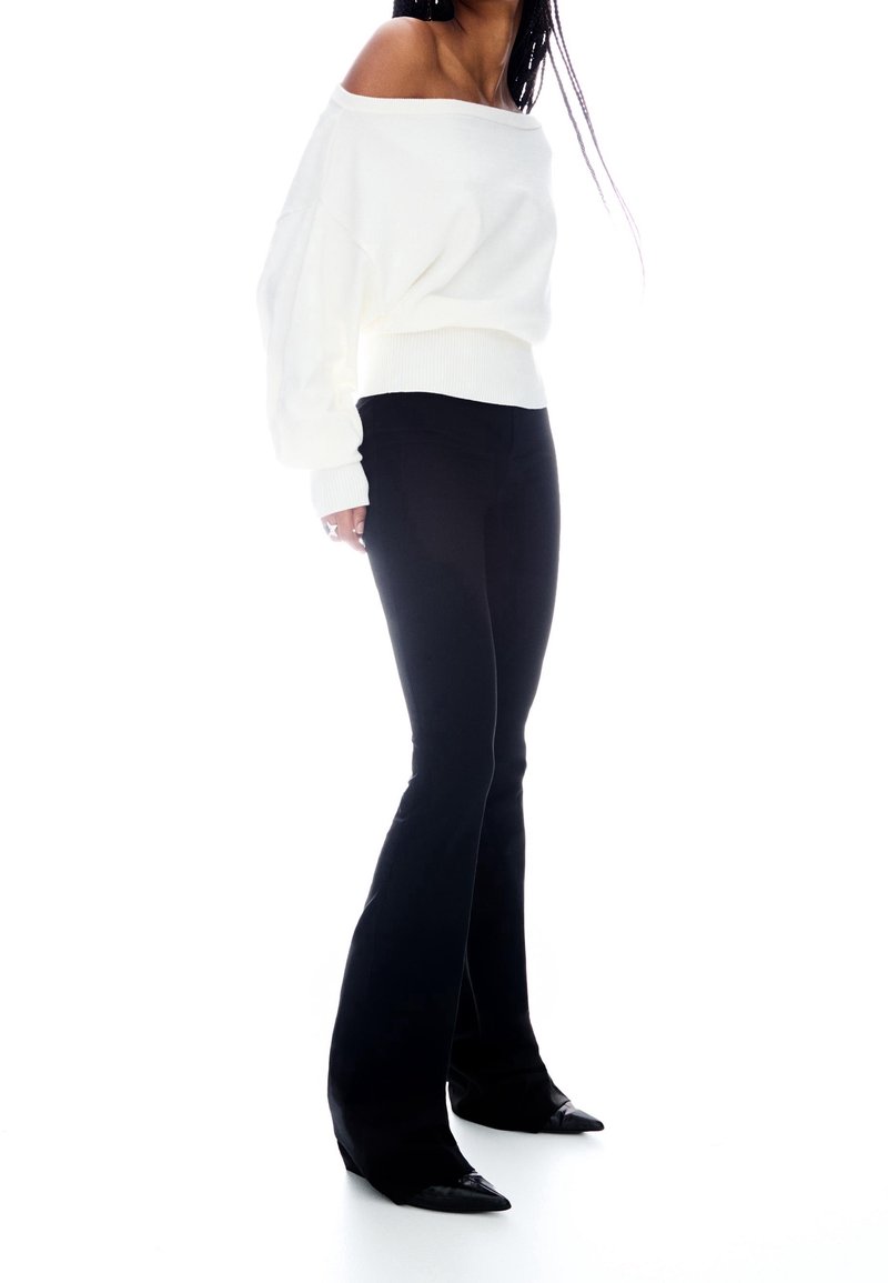 Femme portant un pull blanc décolleté aux épaules, un pantalon noir évasé ajusté et des chaussures noires à bout pointu, debout devant un fond blanc.