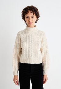 Pulover crem din tricot cu model cable-knit, cu guler înalt și mâneci balon, având modele texturate. Asortat cu pantaloni negri din catifea răsucită.