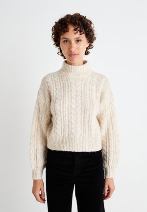 Cream cable-knit sweater with a high collar and balloon sleeves, featuring textured patterns. Paired with black corduroy pants.