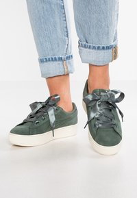 Grüne Wildleder-Sneaker mit einer Plateausohle, ausgestattet mit breiten Satin-Schnürsenkeln und seitlichen Details. Getragen mit hochgekrempelten hellblauen Jeans.
