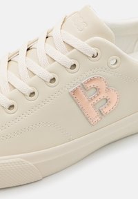 Baskets couleur crème avec lacets texturés, semelle beige et logo "B" en or rose brodé sur le côté près des œillets.