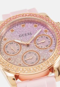 Montre dorée avec un bracelet en silicone rose. Présente un cadran rose pailleté, trois sous-cadrans et une lunette ornée de cristaux roses.