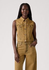 Dzeltena denima bez piedurknēm veste ar apkakli, priekšējiem pogām un krūšu kabatām. Apvienota ar saderīgām augstām biksēm. Smalka tekstūra un krāsas variācija.