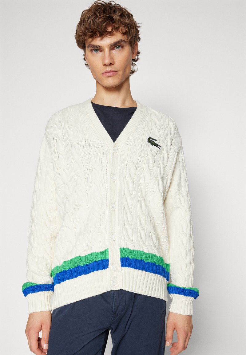 Un jeune homme avec des cheveux bouclés porte un cardigan en maille blanche avec des rayures vertes et bleues, boutonné sur une chemise foncée et un pantalon sombre.