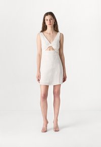 Leikkausmekko vaaleasta beige-linosta, jossa on syvä V-mallinen kaula-aukko ja kierretty etuosa. Tasainen rakenne, istuva siluetti ja polven pituinen muotoilu.