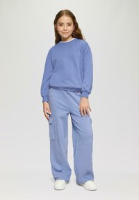 Svetlo modrá mikina s rebrovanými manžetami, v kombinácii s rovnako svetlo modrými cargo nohavicami s bočnými vreckami. Biely tenisky dotvárajú celý outfit.