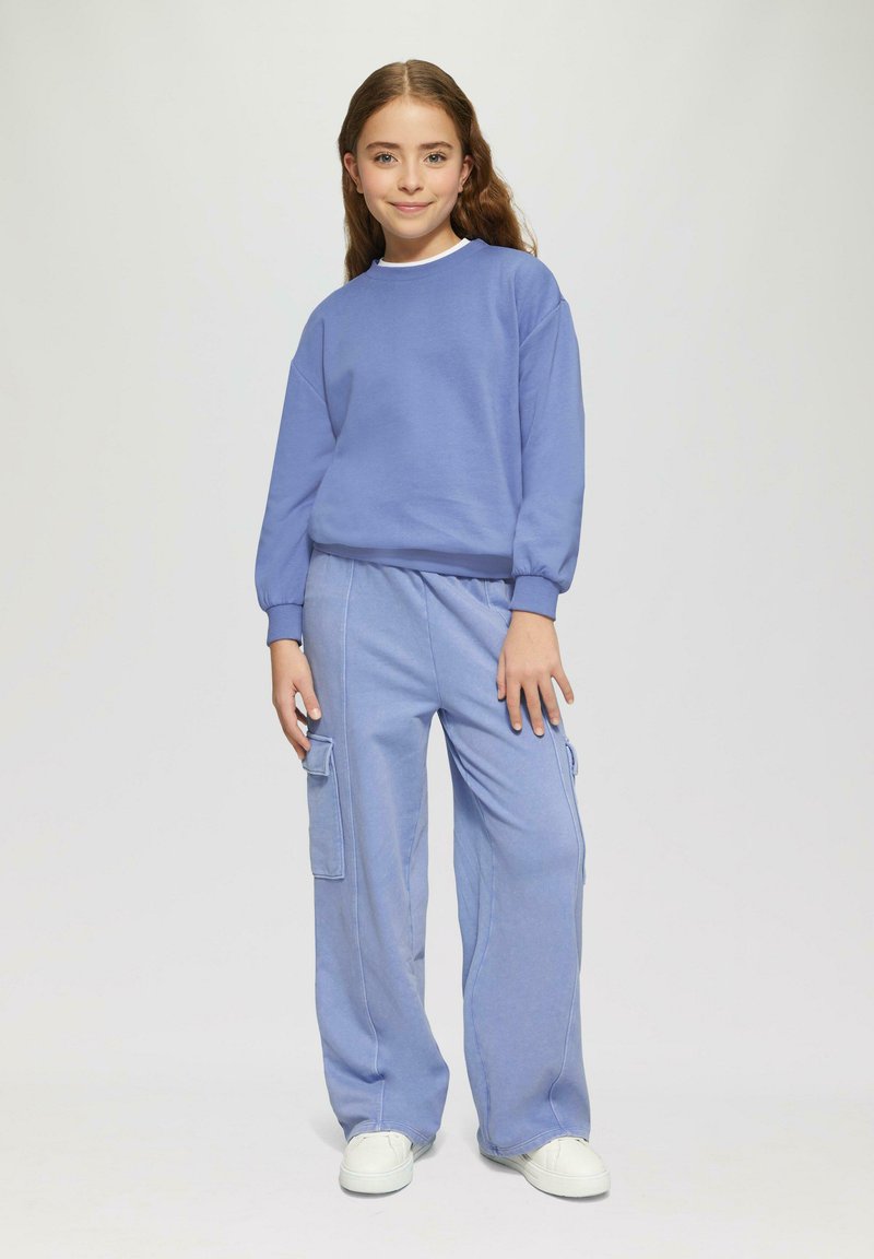 Svetlo modrá mikina s rebrovanými manžetami, v kombinácii s rovnako svetlo modrými cargo nohavicami s bočnými vreckami. Biely tenisky dotvárajú celý outfit.