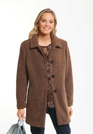 Manteau en polaire marron avec un col, des boutons sur le devant, des poches latérales et une surface texturée. Le mannequin tient un sac à main bleu clair.