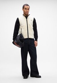 Ανδρικό γιλέκο φλις με φερμουάρ σε κρεμ απόχρωση και μαύρα μανίκια, συνδυασμένο με σκούρα φαρδιά τζιν και μαύρη τσάντα. Απαλή υφή, κα casual σχεδίαση.