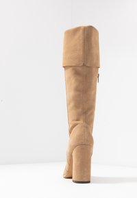 Botte hauteur genou en daim beige avec un talon épais, dotée d'un design à rabat en haut et d'une fermeture éclair latérale.