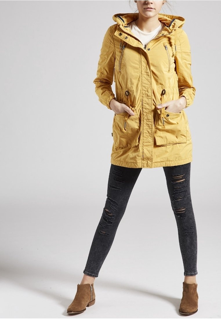 Femme portant une veste jaune à capuche, un jean skinny noir déchiré et des bottines marron, debout les mains dans les poches de la veste.