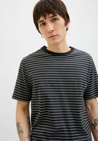 Sortstribet t-shirt med vandrette hvide striber, korte ærmer, rund hals og en glat stofstruktur. Fremhævet med synlige tatoveringer.