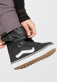 Zapatilla negra con acentos blancos y una suela de goma texturizada. La persona está ajustando el dobladillo de un pantalón sobre el zapato.