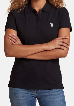 Personne portant un polo noir à manches courtes avec un petit logo blanc brodé, les bras croisés, porté avec un jean bleu.