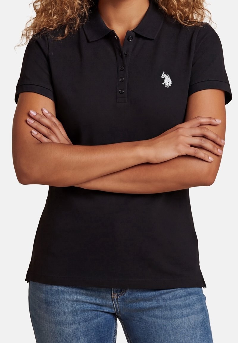 Personne portant un polo noir à manches courtes avec un petit logo blanc brodé, les bras croisés, porté avec un jean bleu.