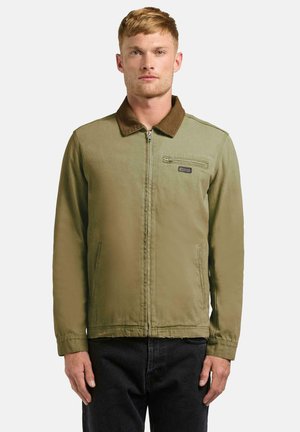 Ung mann iført en olivengrønn jakke med glidelås, brun krage og svarte bukser, står mot en ensfarget lys grå bakgrunn.