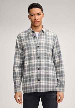 Mann trägt ein grau-weiß kariertes Hemd über einem weißen T-Shirt, steht vor einem schlichten hellen Hintergrund.