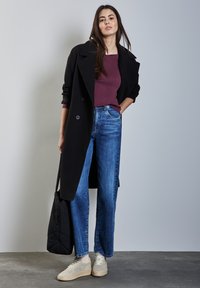 Schwarzer langer Mantel über einem kastanienbraunen Oberteil, kombiniert mit blauen weit geschnittenen Jeans und hellbeige Sneakers. Eine schwarze Tote-Bag hängt über einer Schulter.