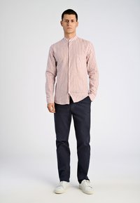 Camicia a righe a maniche lunghe in rosso chiaro e bianco, con colletto, bottoni frontali e taschino sul petto, abbinata a pantaloni blu navy e sneakers bianche.
