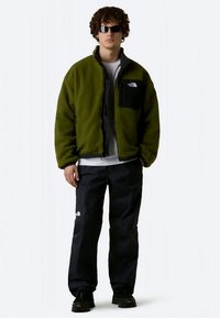 Uomo con occhiali da sole scuri, giacca in pile verde, pantaloni neri e scarpe nere, in piedi contro uno sfondo neutro.