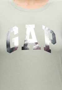 Gros plan d’une chemise vert clair avec de grandes lettres argentées brillantes formant le mot GAP sur la poitrine.