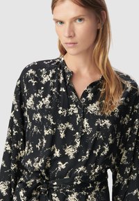 Czarna bluzka w kwiatowy wzór z długimi rękawami, zapięciem na guziki i wiązaniem w talii. Materiał ma gładką fakturę z kremowymi akcentami.